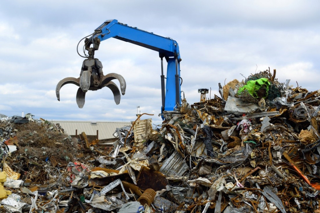 A scrap yard, with piles of scrap metal around. A large blue arm of a crane has a claw on the end and is hovering above the scrap metal.